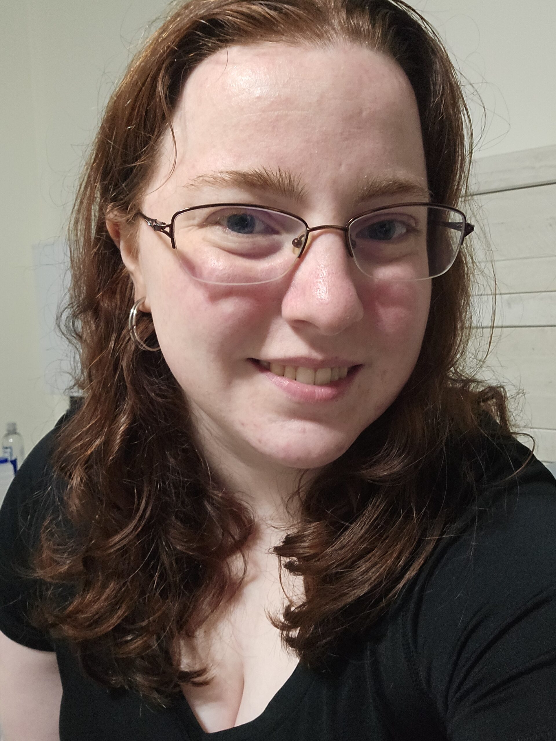 A selfie of Rosie Caudle. Red wavy hair, fair skin, copper frame glasses, black shirt.